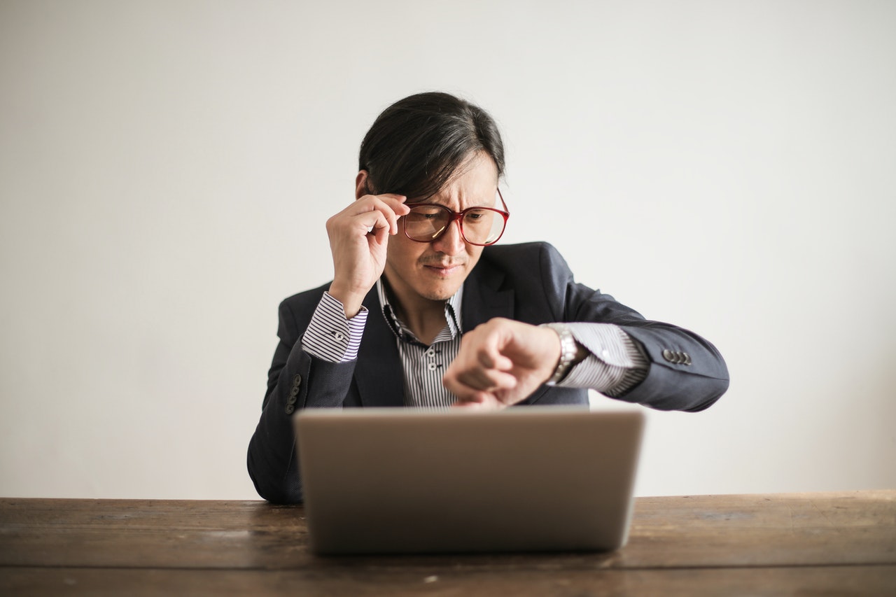 Hombre sentado al frente de un computador portatil y mirando su reloj.